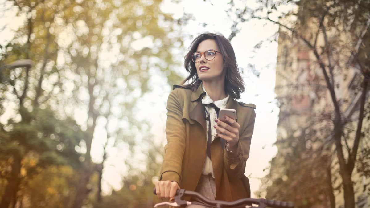 Stylish woman riding a bike, holding a smartphone on a sunny autumn day outdoors.